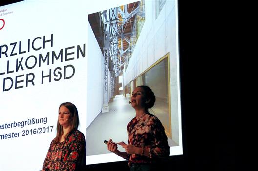 Die Studentinnen des Fachbereiches Wirtschaft, Nicola Machaczek and Carla Reichle, stehen auf der Bühne der Mitsubishi Electric Hall. Im Hintergrund zeigt eine Präsentation das Logo der Hochschule, den Schriftzug "Herzlich willkommen an der HSD" und ein Bild des Interieurs des Gebäudes der Campus IT.